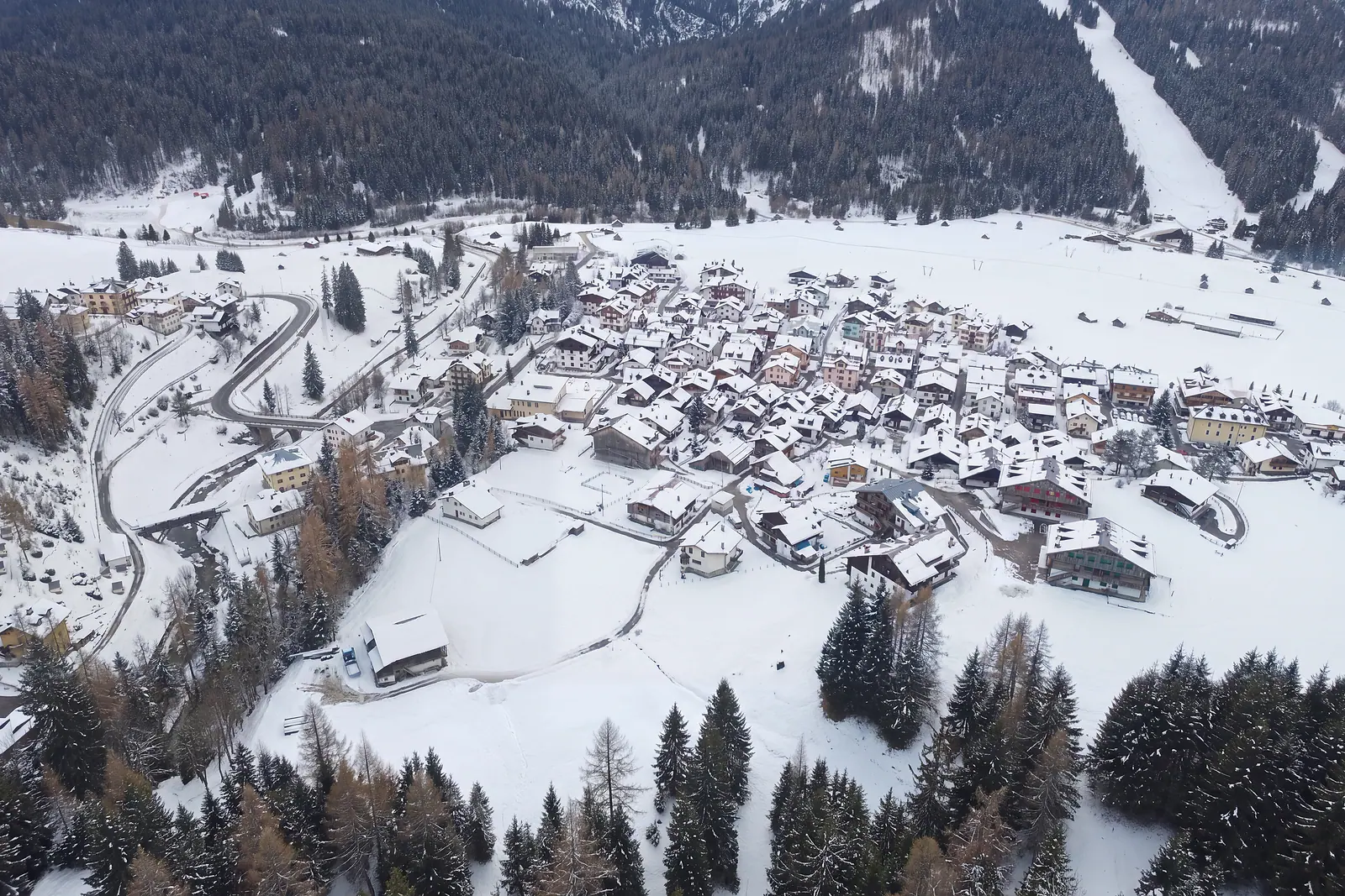 Die ganze Borgata Bach in einer Winterfassung, schräg von Nordwesten gesehen: verschneite Dächer, weiße Wiesen, links die Schlucht des Bachs mit den Kehren der Straße. Mit KI aus einem echten Frühjahrsfoto bearbeitetes Bild.