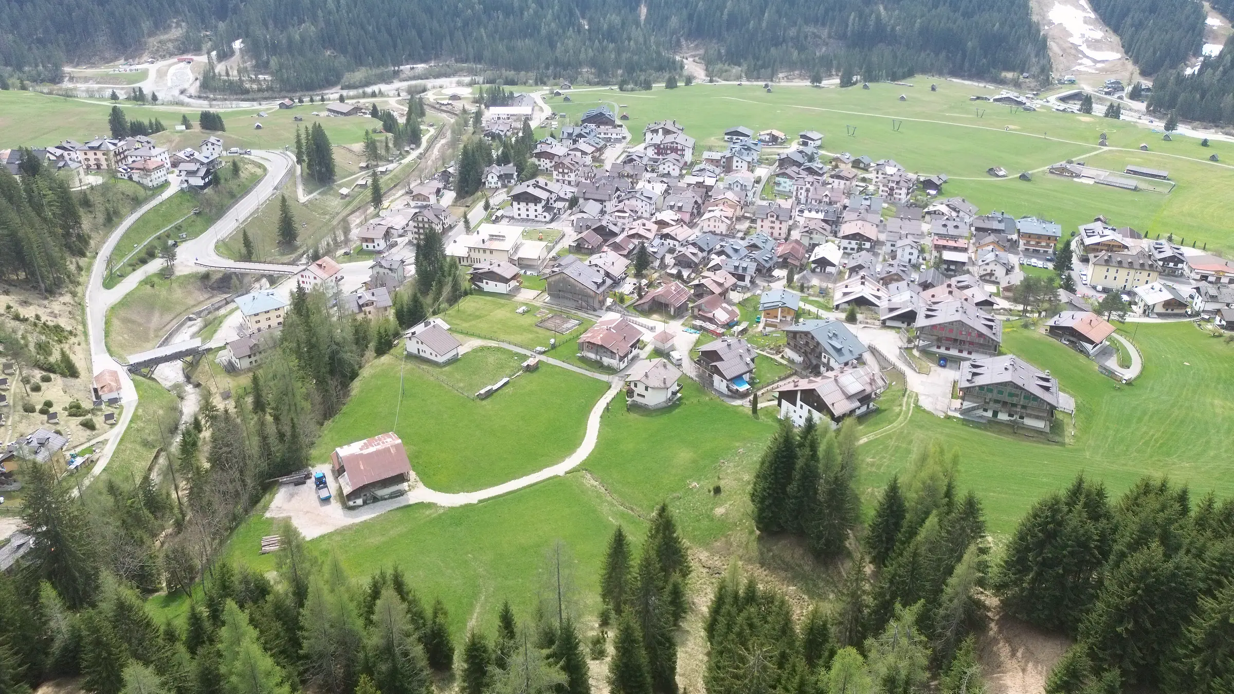 Die ganze Borgata Bach, schräg von Nordwesten gesehen: in der Mitte die dicht stehenden dunklen Dächer, ringsum Wiesen, links die Bachschlucht mit den Kehren der Straße.
