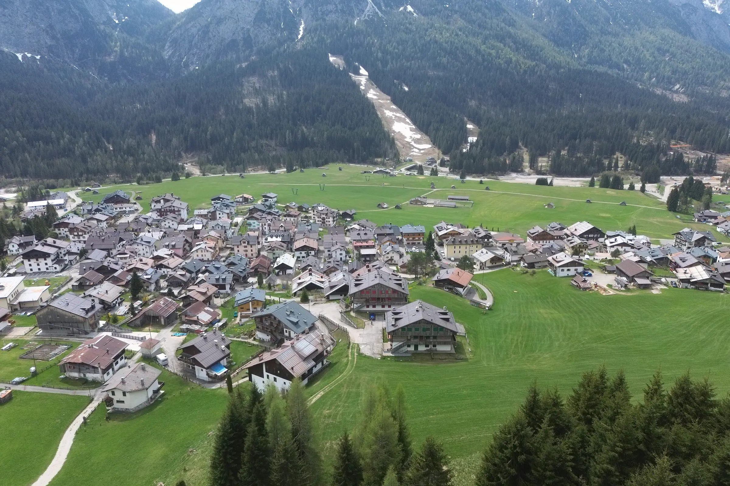 Die Borgata Bach von oben mit der Wiesenebene und den verstreuten Heustadeln; jenseits des Piave der Wald und die Skipiste mit den letzten Schneeflecken.