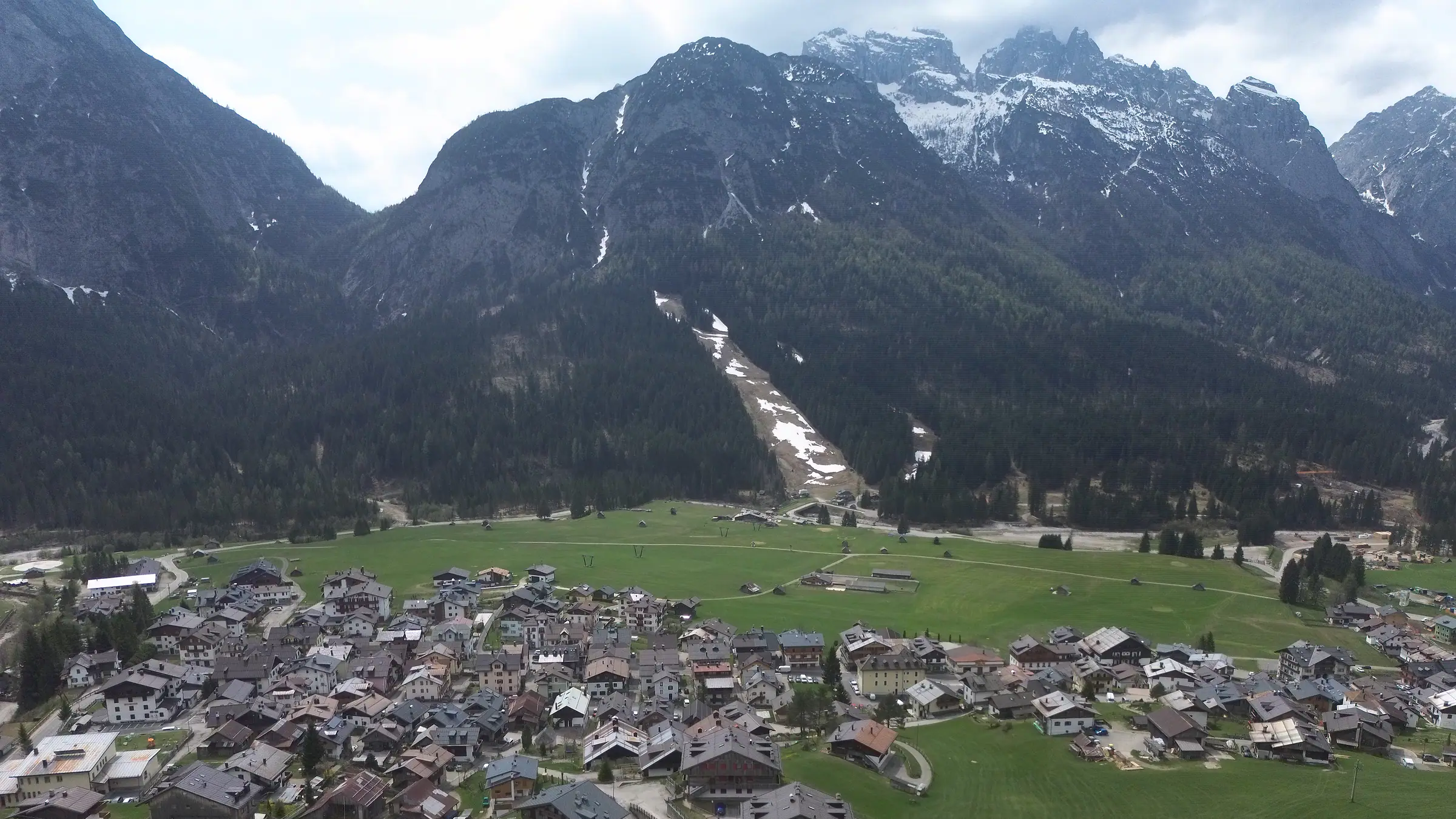 Die Borgata Bach von oben, die Häuser dicht zwischen den Wiesen; über dem Wald die hellen Felswände und der letzte Schnee der Clap-Gruppe.