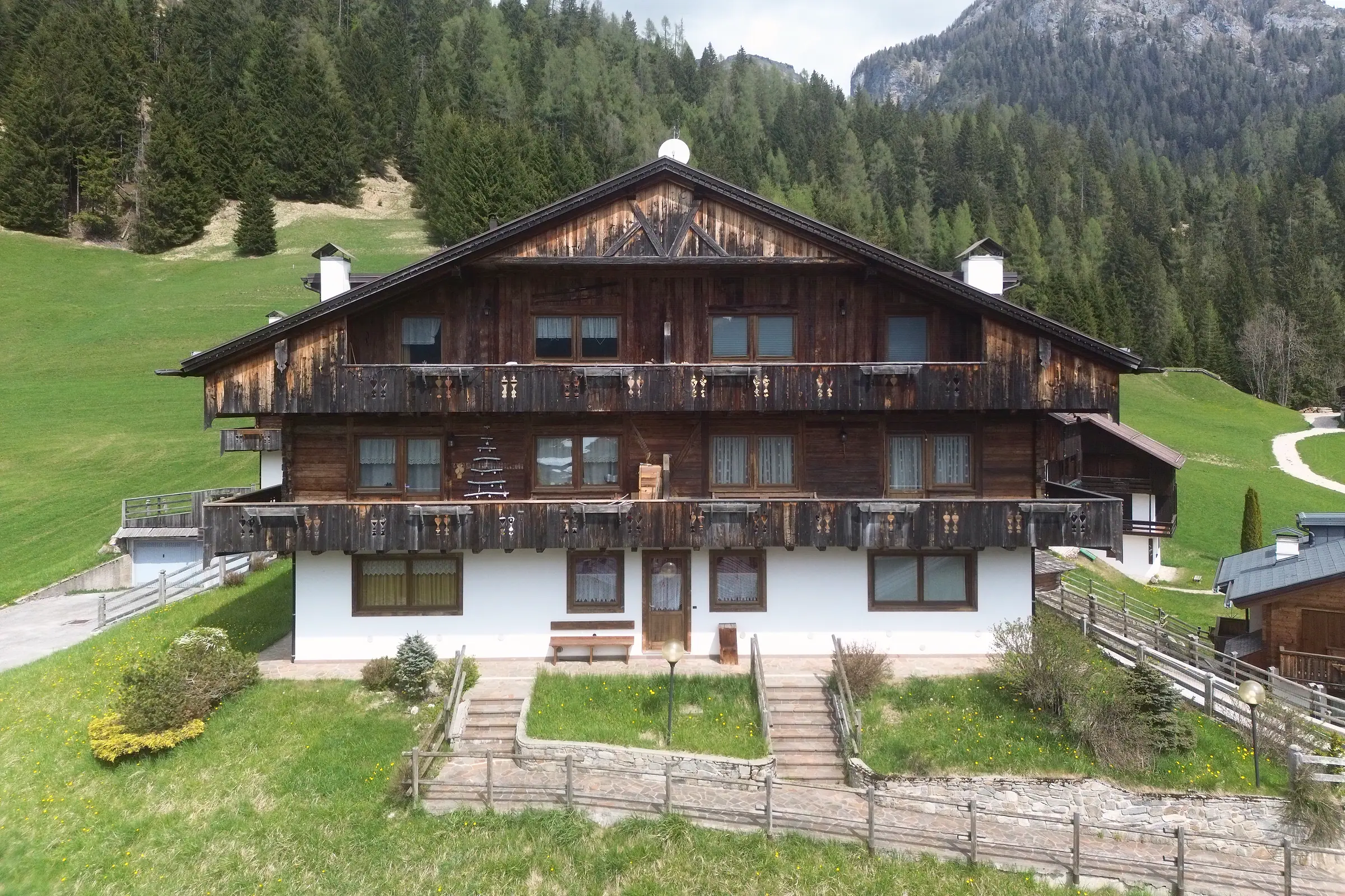 Ein Haus in der Borgata Bach von vorn: weiß verputztes Erdgeschoss, zwei Reihen geschnitzter dunkler Holzbalkone, verbretterter Giebel und Satteldach.