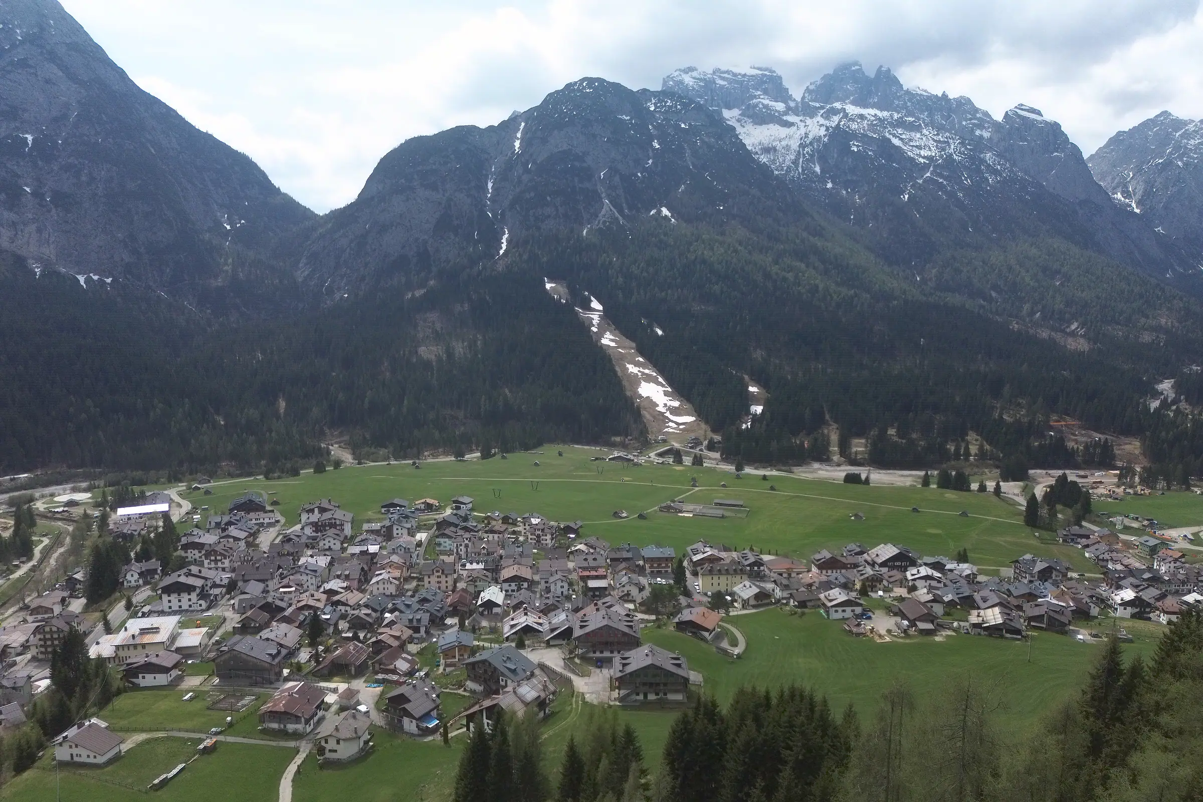 La conca di Sappada vista dall'alto sopra Borgata Bach: i tetti della borgata, la piana a prato, il bosco e in fondo le cime ancora innevate del gruppo dei Clap.