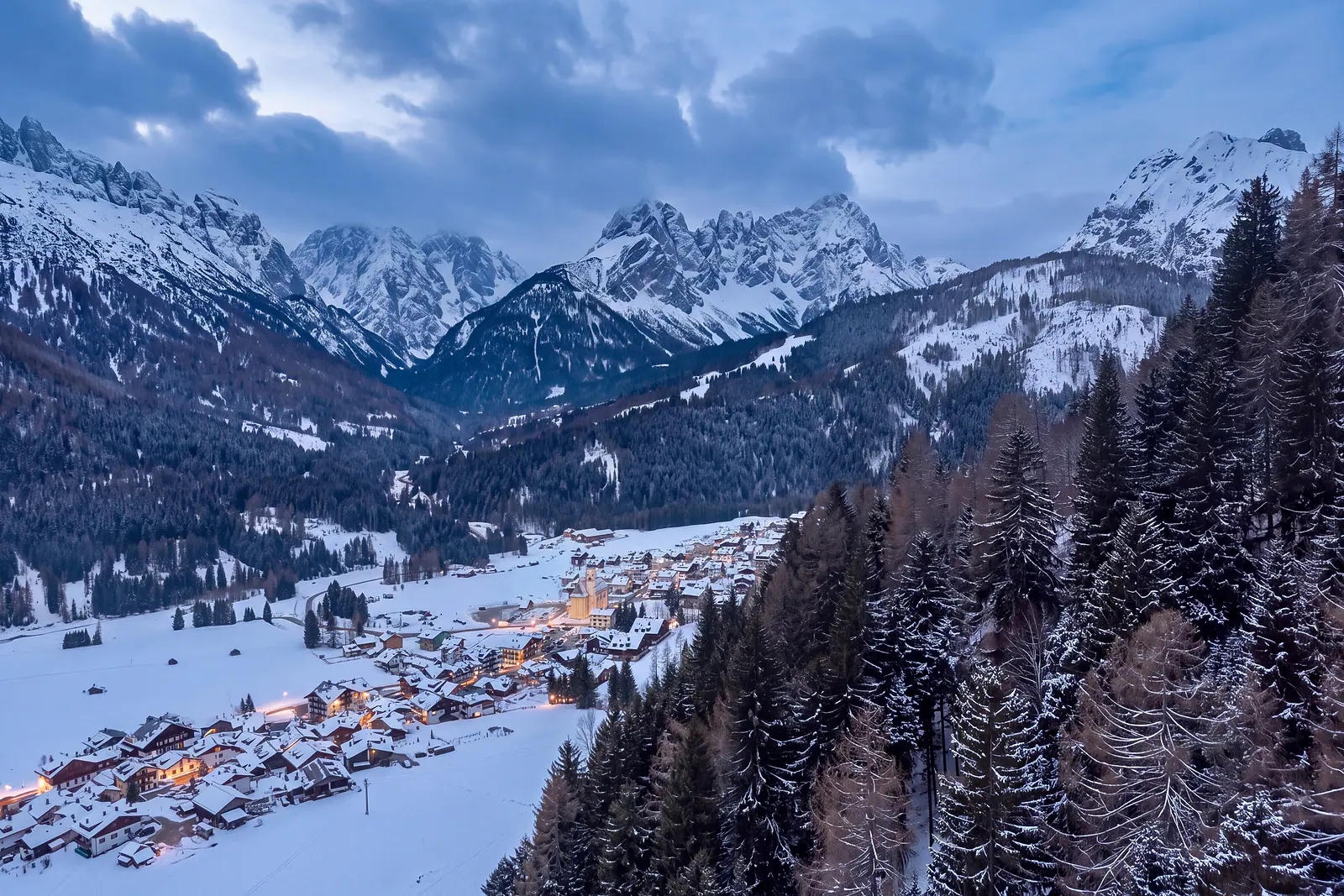 Pill and Granvilla on a simulated winter evening: snow on roofs and meadows, a dark blue sky, windows and street lamps lit among the houses, the parish church with its bell tower and the ridges of the Terze in the background. Image reworked with AI from a real photograph taken in daylight, in spring.