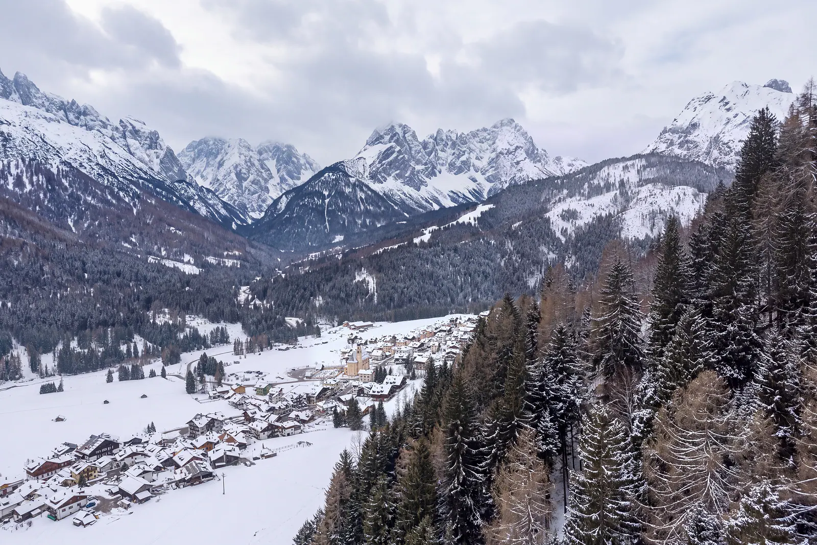 The valley towards the west in a winter version: in the foreground the houses of Pill under snow, in the centre Granvilla with the bell tower of the parish church, in the background the white ridges of the Terze. Image reworked with AI from a real spring photograph.