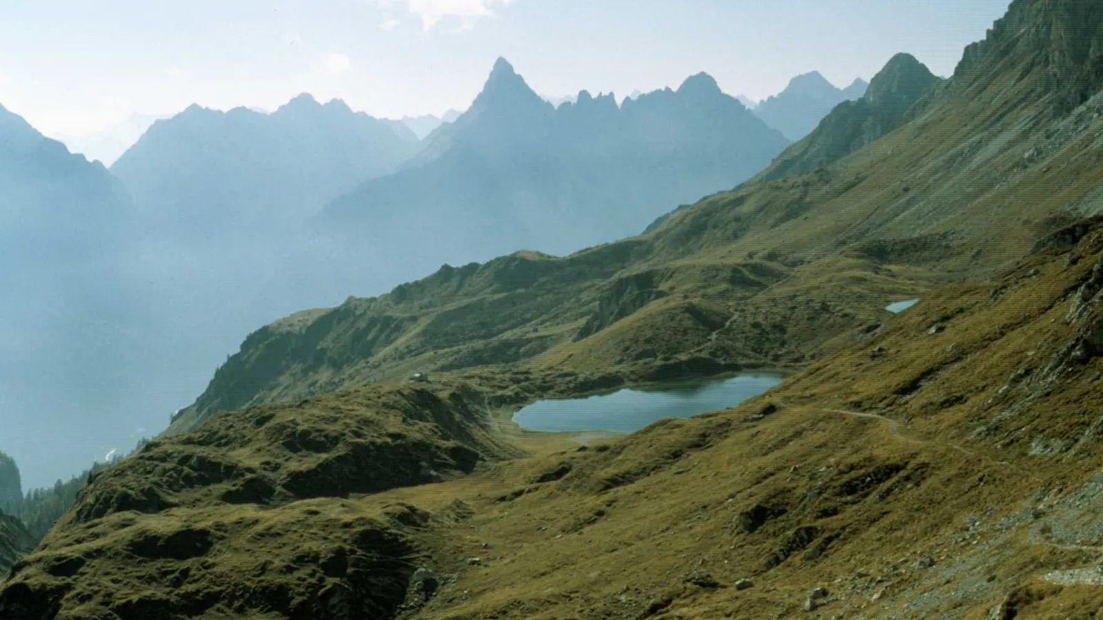 One of the Olbe lakes, above Sappada: a small alpine lake in a hollow of pastures, with the outline of the Terza Grande behind in the haze.