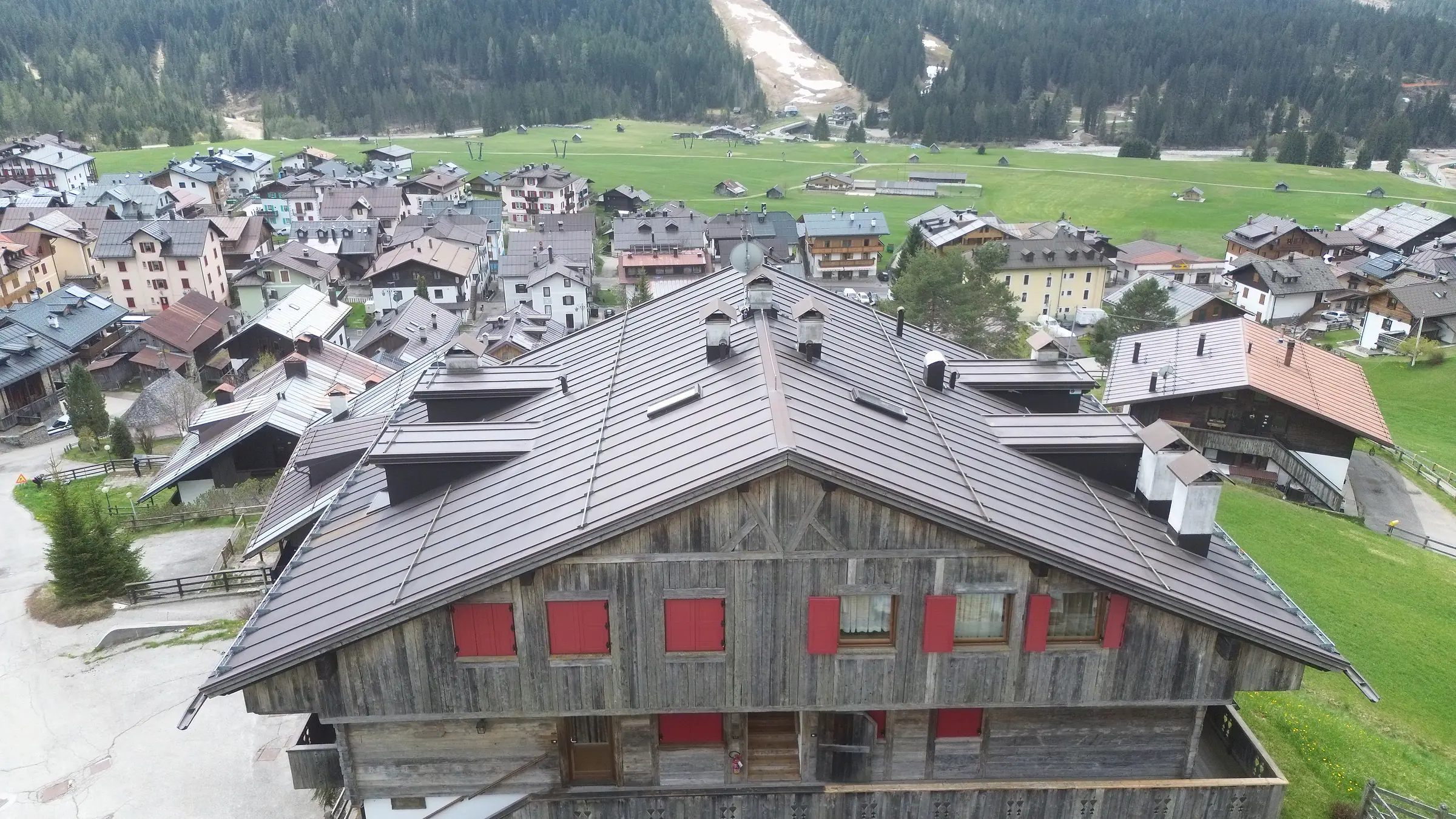 The roof and weathered grey timber gable of a house in Bach, with red shutters; behind it, the roofs of the hamlet, the meadow plain and the ski run beyond the Piave.