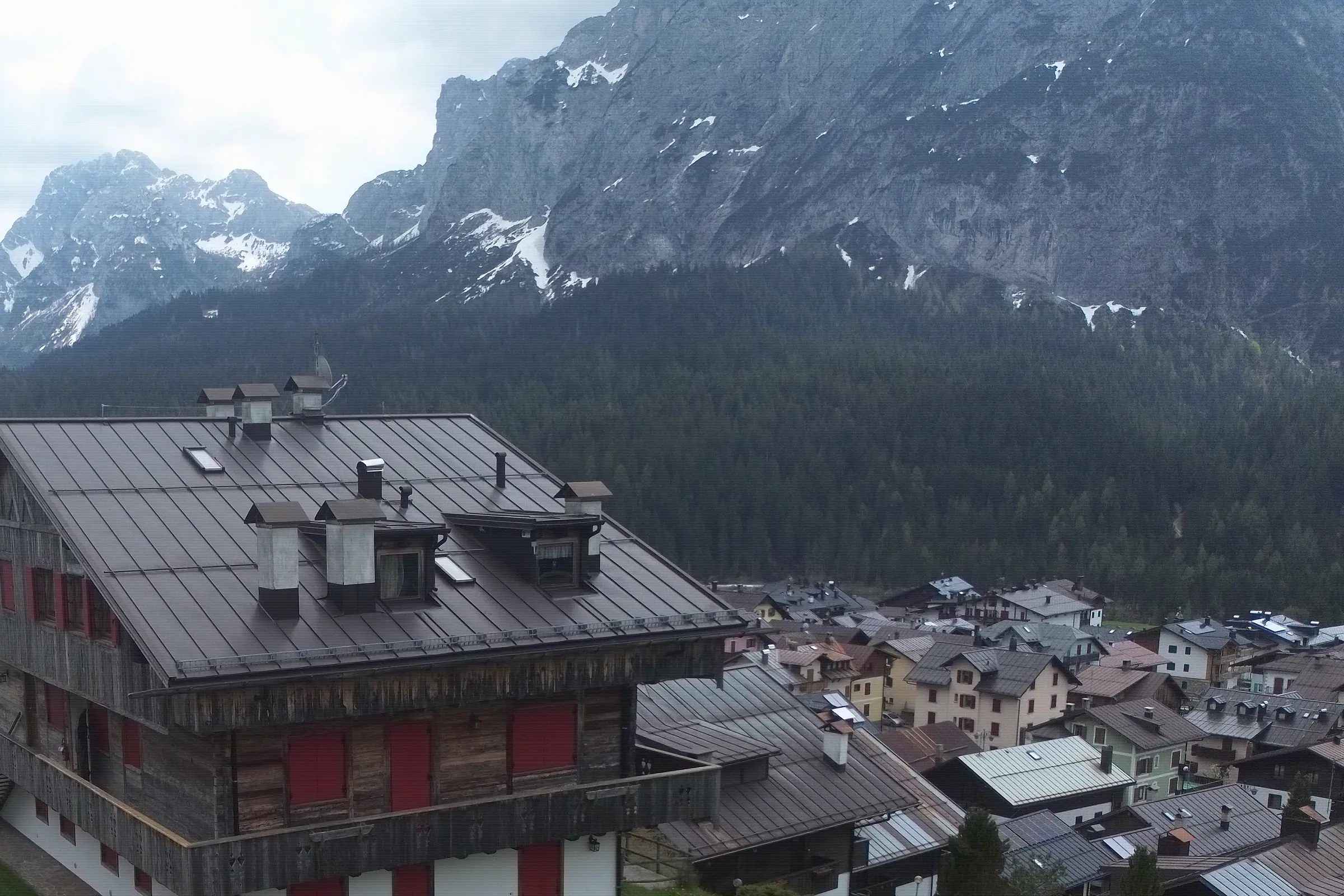 Ein dunkles Blechdach mit Kaminen und Gauben, dahinter die Felswand des Monte Siera mit den letzten Schneezungen; weiter entfernt der Monte Cimon.