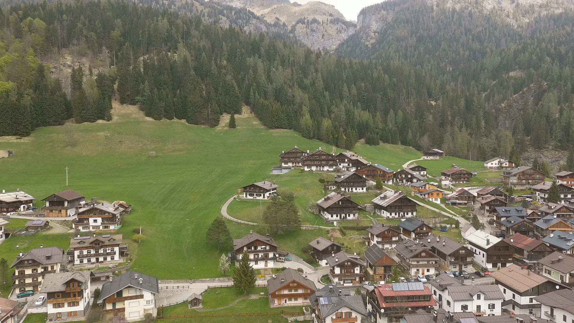 Ripresa dal drone verso nord: le case in legno della parte alta di Borgata Bach, il prato che sale fino al bosco di abeti e larici e, sopra, le rocce chiare del versante al sole.