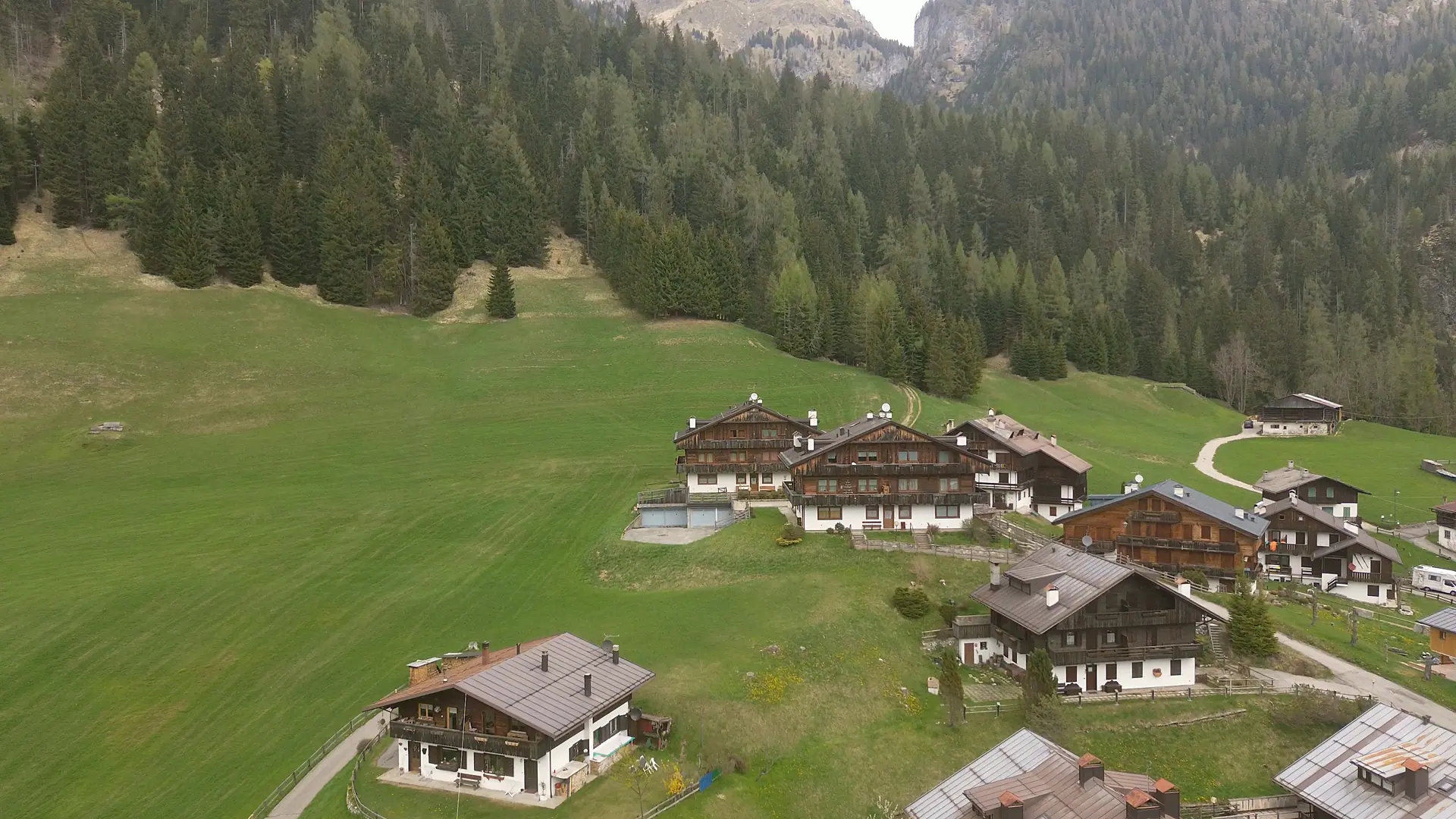 Posnetek z dronom, ki se približuje lesenim hišam zadnje vrste zaselka Bach: belo ometano pritličje, zgornja nadstropja iz temnega lesa z balkoni, okrog travnik in zadaj gozd.