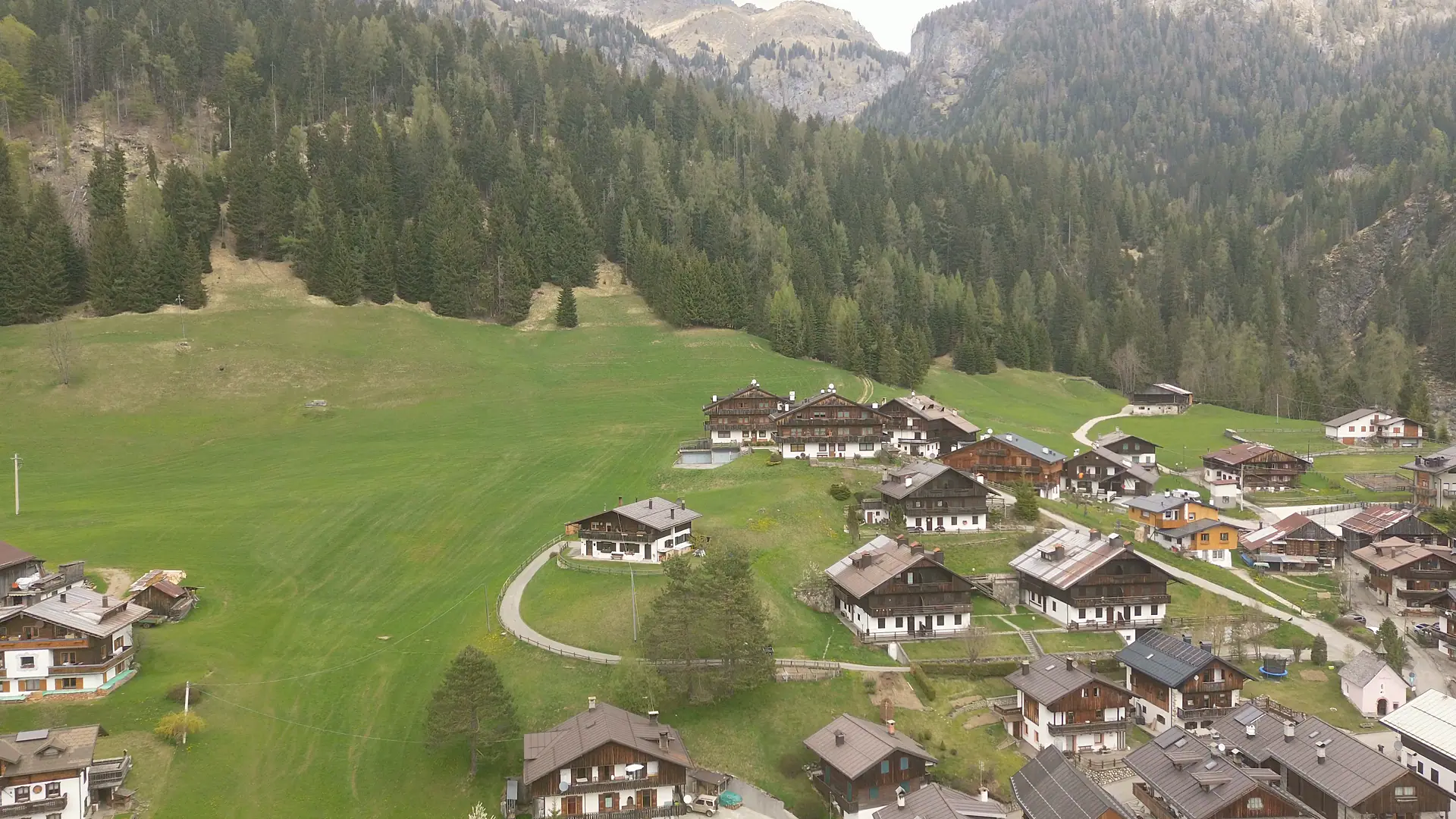 Drone footage looking north: the cluster of timber-and-render houses in the upper part of Borgata Bach, gathered on the sloping meadow below the edge of the forest.