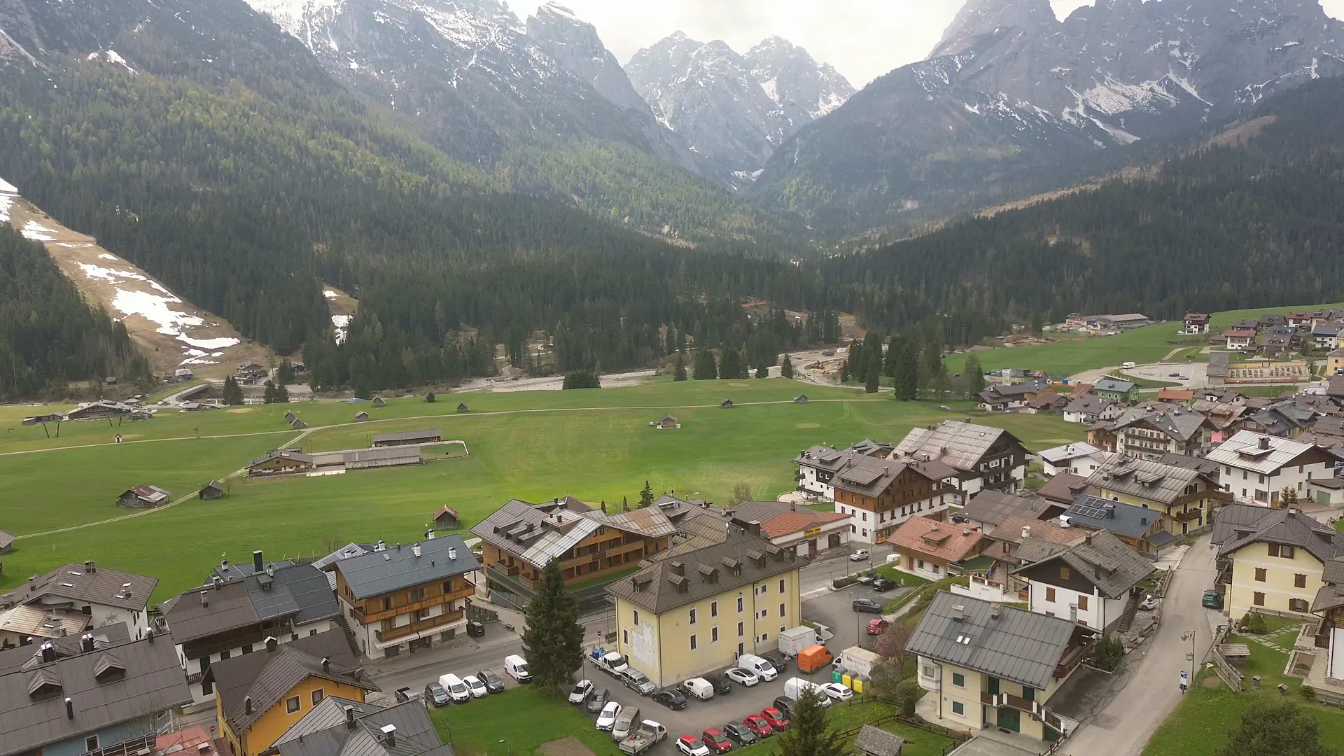 Posnetek z dronom nad zaselkom Bach: strehe zaselka, travnata ravnica s seniki in smučišče onkraj Piave; kamera se počasi obrne proti zahodu do Granville pod grebeni skupine Terze.
