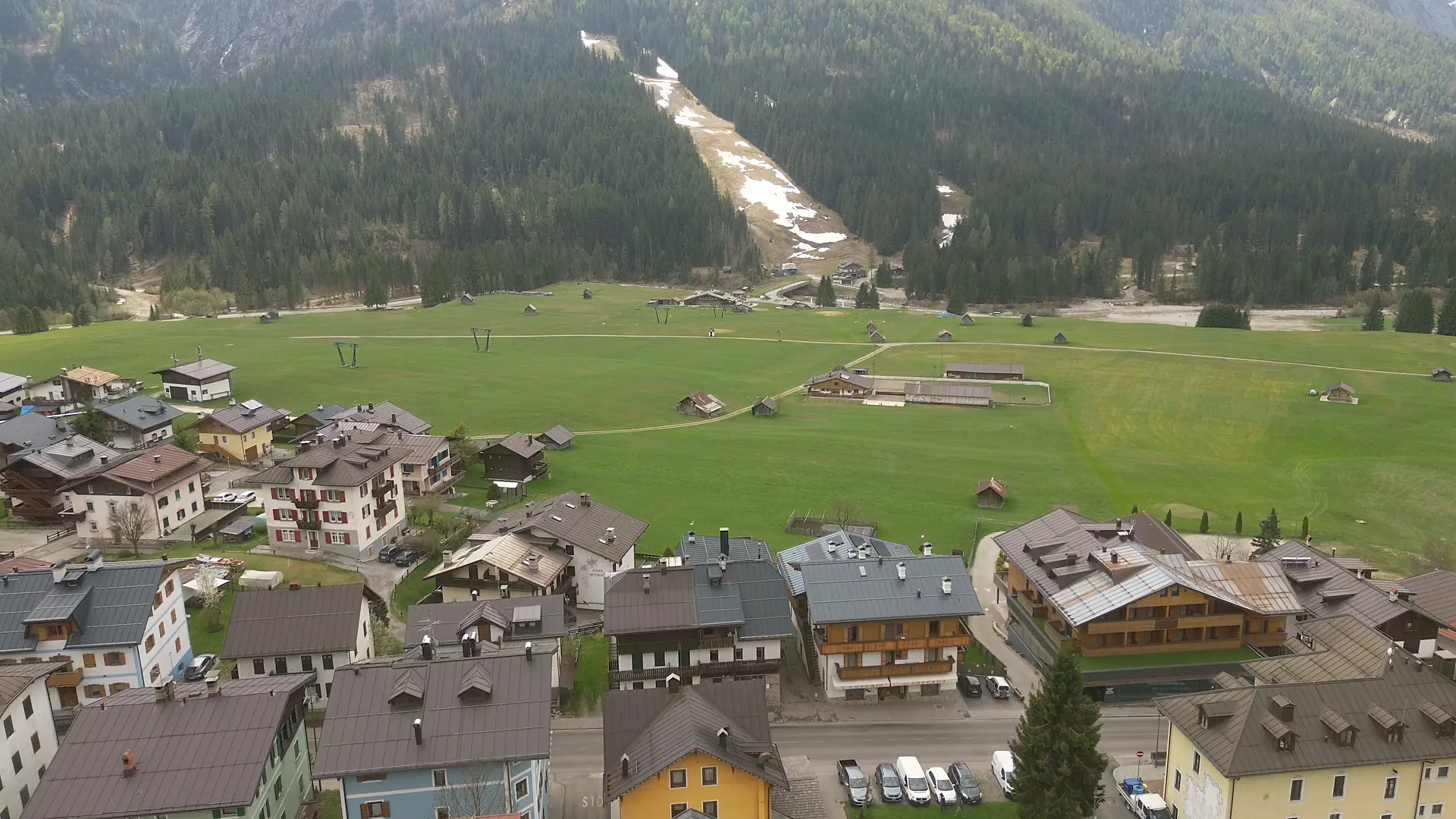 Drone footage over the edge of Borgata Bach: the last houses, some recent and timber-clad, then the meadow plain with its small hay barns and the ski run beyond the Piave.