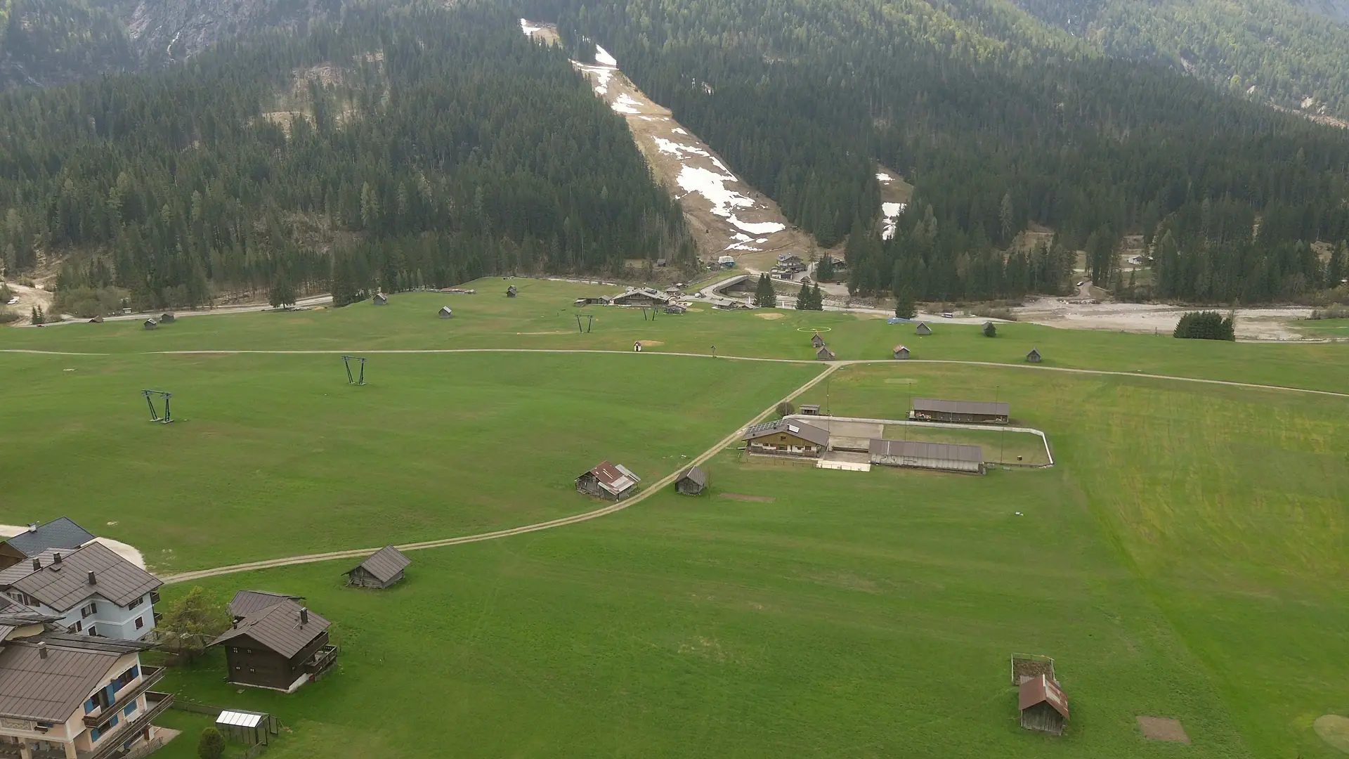 Drone footage above the plain of Sappada: hay meadow dotted with small timber barns, a gravel track, in the background the bed of the Piave, the forest and the ski run with its last patches of snow.