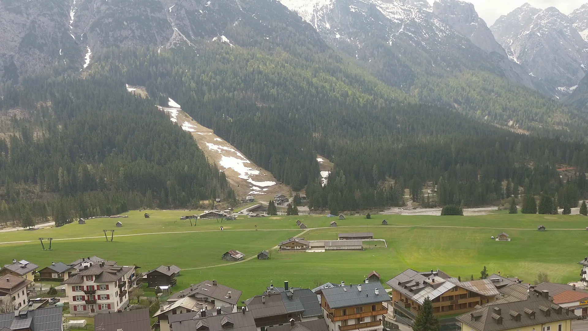 Ripresa dal drone verso sud: la piana a prato coi fienili, oltre il Piave la pista da sci che sale nel bosco e, in alto, le cime ancora innevate; in basso i tetti delle ultime case di Borgata Bach.