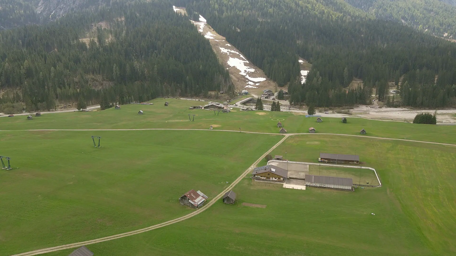 Drohnenaufnahme über der Ebene von Sappada: die Mähwiese mit einem Schotterweg, die Heustadel, das helle Kiesbett des Piave und dahinter die Skipiste im Wald.