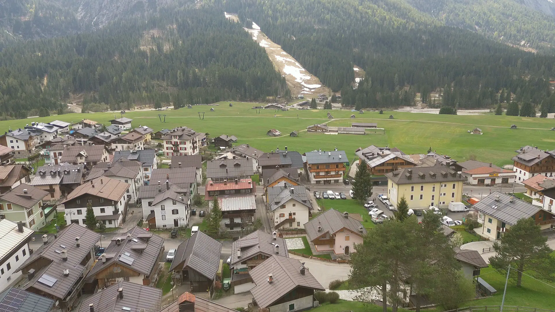 Drohnenaufnahme über den Dächern der Borgata Bach: dicht stehende zwei- und dreigeschossige Häuser mit dunklen Blechdächern, dahinter die Wiesenebene und die Skipiste im Wald.
