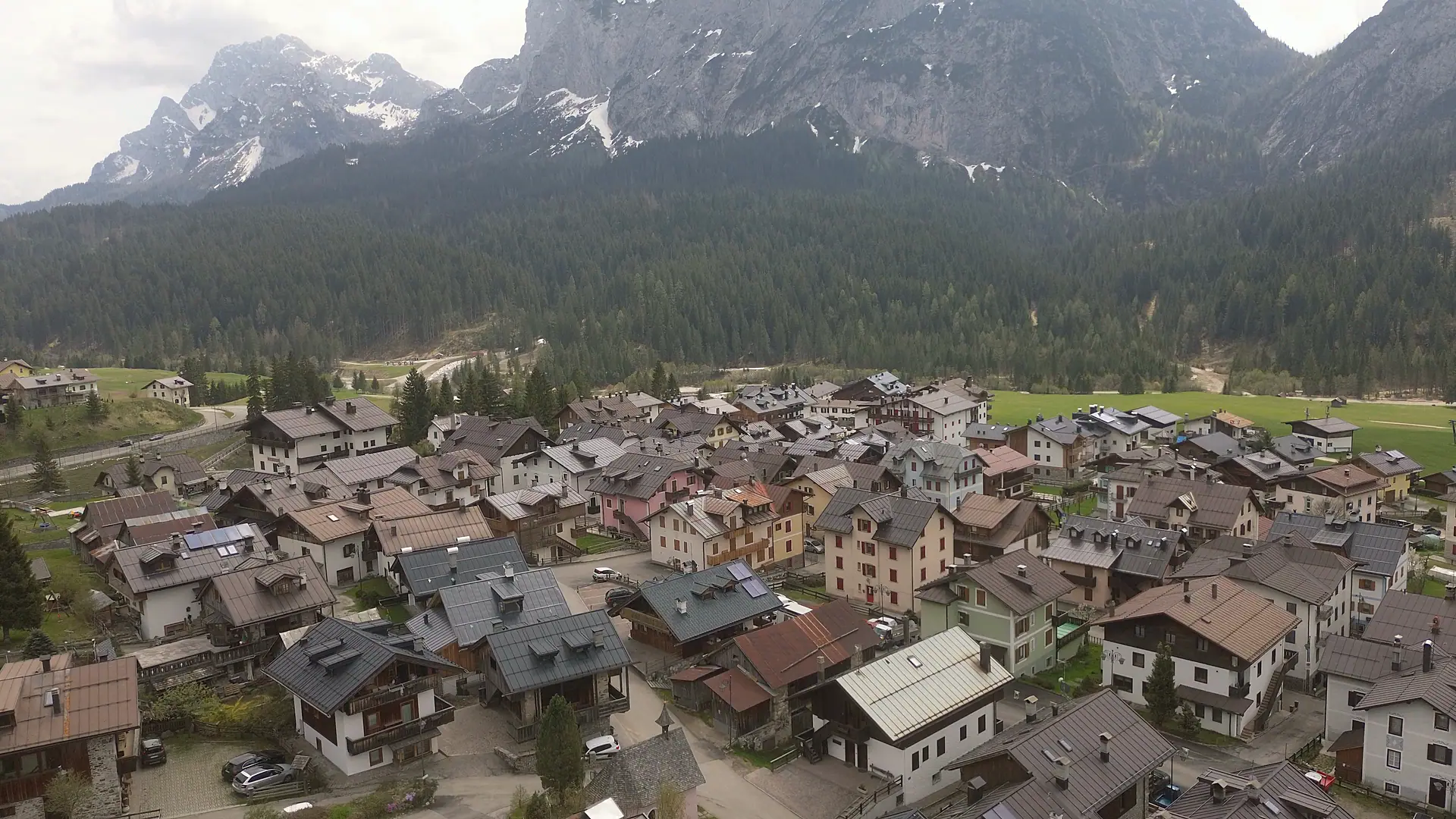 Drone footage above Borgata Bach looking south-east: the closely set roofs of the hamlet, the spruce forest on the slope opposite and, above, the rock walls with the last tongues of snow.